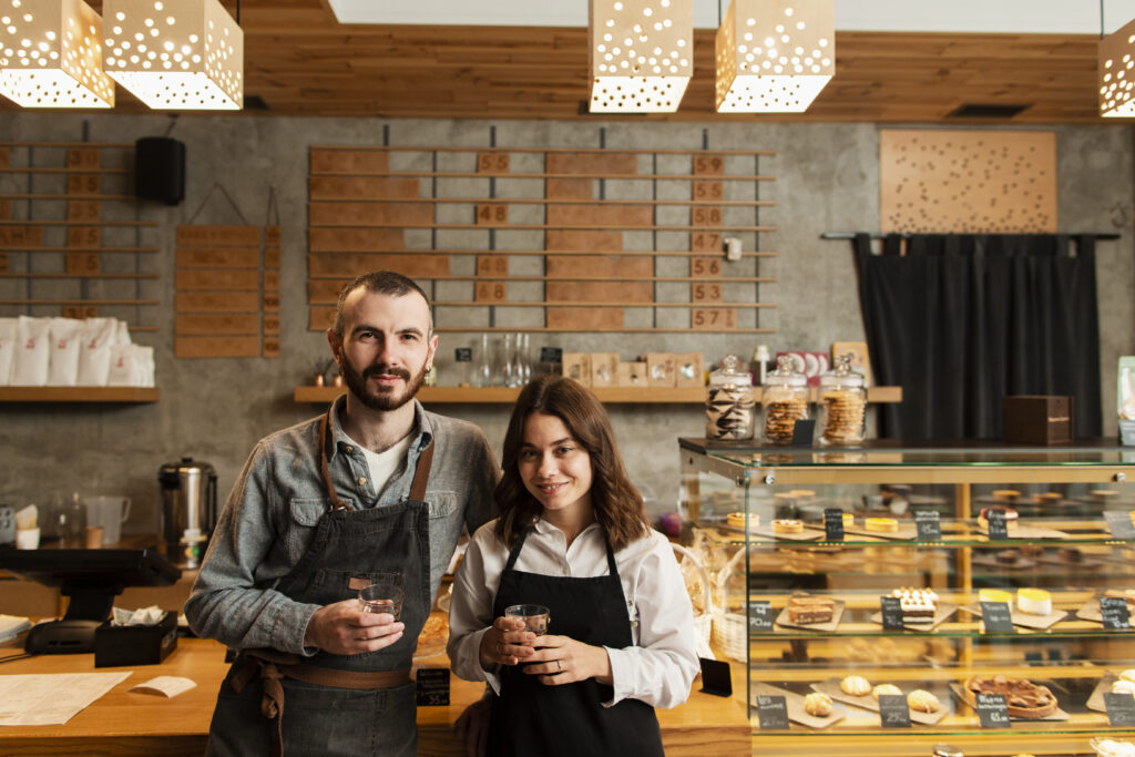 Two small business owners standing behind the counter of their cozy bakery, smiling and holding cups, with various pastries displayed in the background.