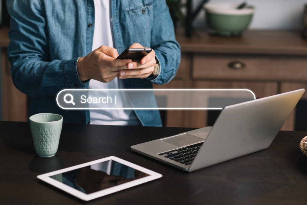 A person using a smartphone at a desk with a laptop and tablet, looking at a search bar, representing North Texas Digital Marketing, a leading SEO company in Dallas.